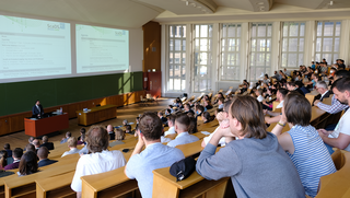 Das Bild zeigt einen Vorlesungssaal. Die Personen sitzen auf einer zur Präsentationsfläche hin abfälligen Sitzstruktur.